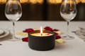 Candle in a black holder on a table with wine glasses and rose petals, blurred lights in the background